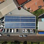 Vista aérea de um prédio comercial com painéis solares instalados no telhado e estacionamento com carros ao redor.