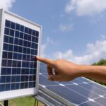 Mão apontando para um painel solar fotovoltaico em um ambiente ao ar livre, com céu azul, nuvens leves e vegetação ao fundo.