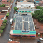 Vista aérea de um supermercado chamado "Stábile" com placas solares no telhado, cercado por casas e árvores em uma área urbana.