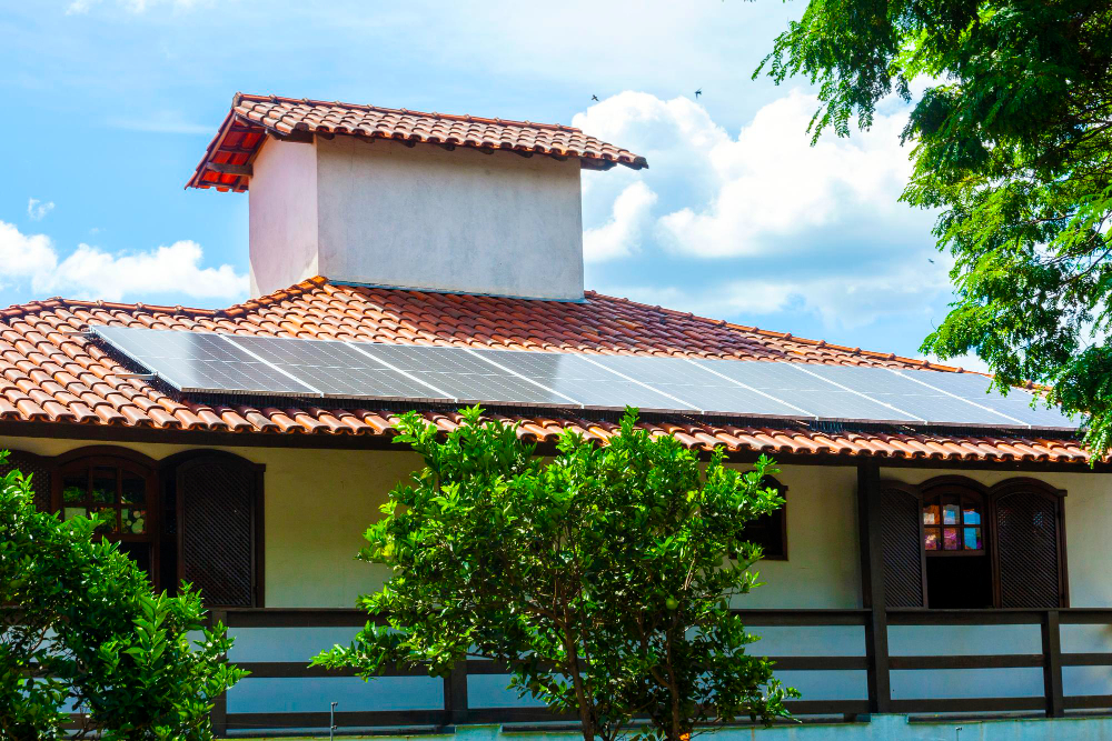 Casa com telhado de telhas cerâmicas equipada com painéis solares, cercada por árvores e iluminada por um céu ensolarado com nuvens.