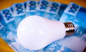 Lâmpada LED branca sobre várias cédulas de 100 reais dispostas em leque, simbolizando o aumento no custo da energia elétrica.