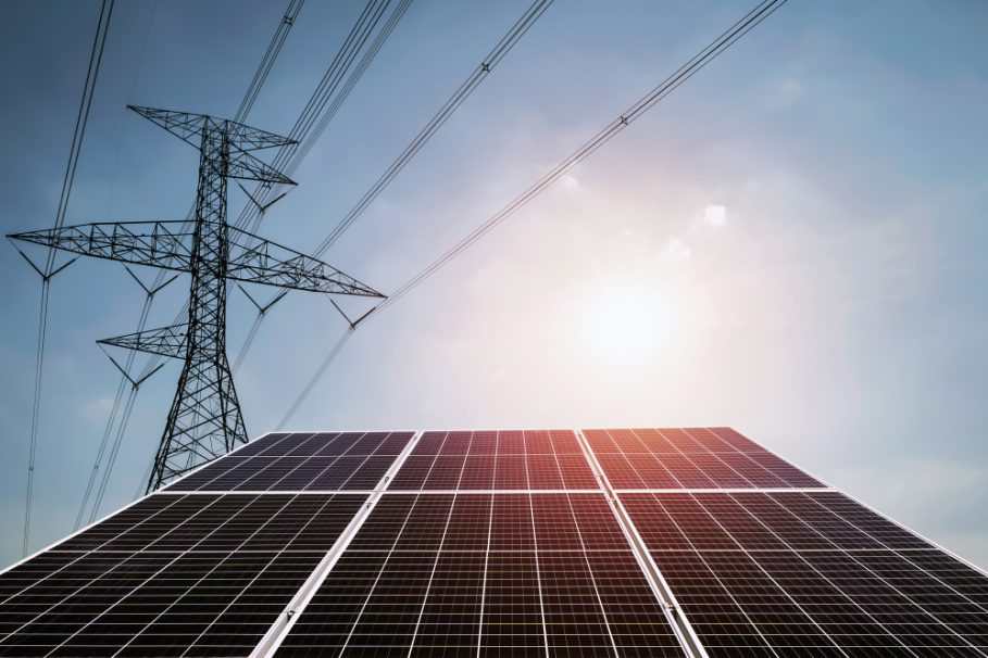 Painéis solares sob luz solar intensa, com torre de transmissão de energia ao fundo e céu azul.