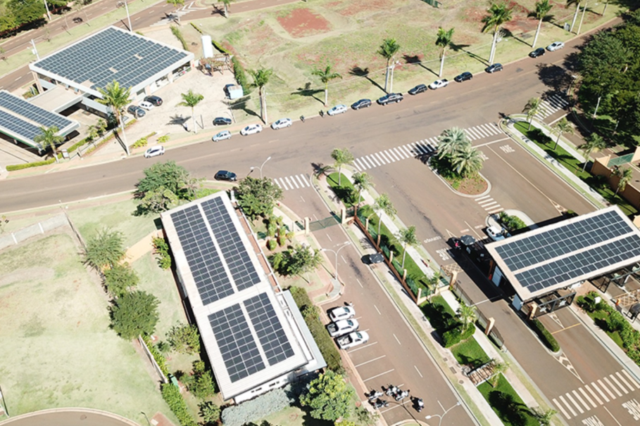 Visão aérea de um complexo urbano planejado, com ruas organizadas, edifícios com painéis solares nos telhados e áreas verdes bem cuidadas, destacando sustentabilidade e infraestrutura moderna.