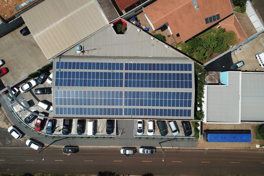 Vista aérea de um prédio comercial com painéis solares instalados no telhado e estacionamento com carros ao redor.