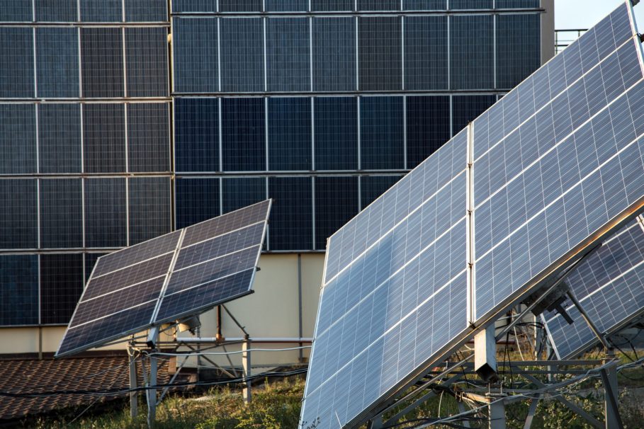 Painéis solares instalados em um campo, com um edifício ao fundo coberto por mais painéis fotovoltaicos.